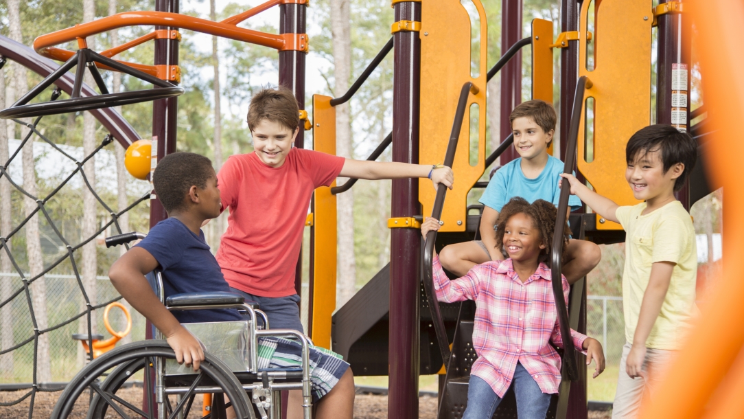 Multi-ethnic group of school children playing on school playground.  African descent, elementary age boy is in a wheelchair.  Other children, play and talk to boy in front of outdoor playground equipment.  Education in USA, exercise themes, differing abilities.