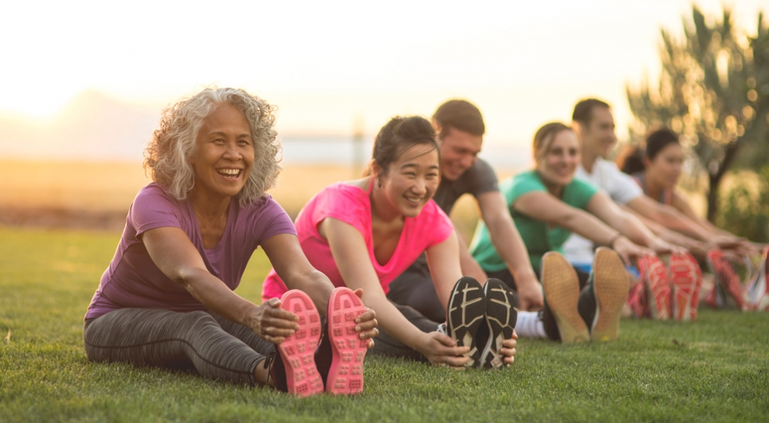 A group of adults attending a fitness class outdoors are doing leg stretches. The participants are arranged in a line. The focus is on a mature ethnic woman who is smiling toward the camera.
